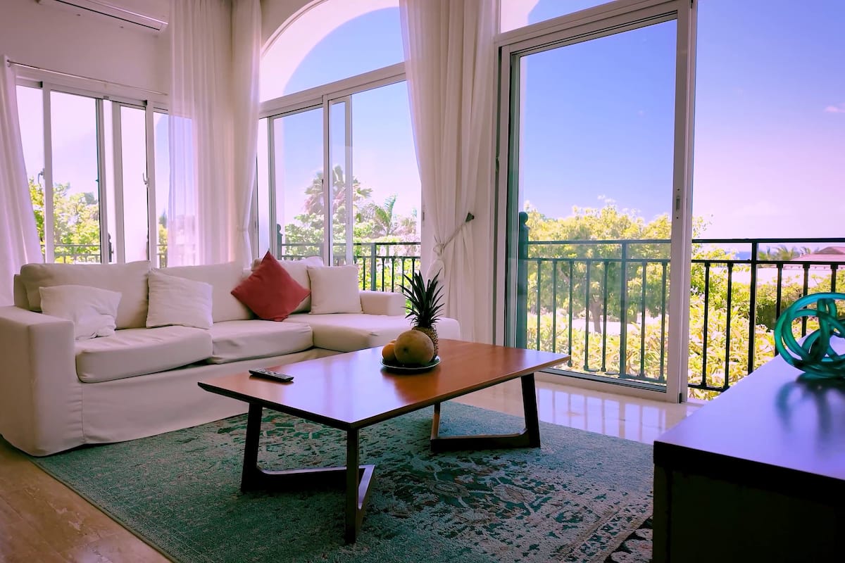 A modern living room with a white sofa, a coffee table with fruits, and large windows offering a view of greenery and a balcony.