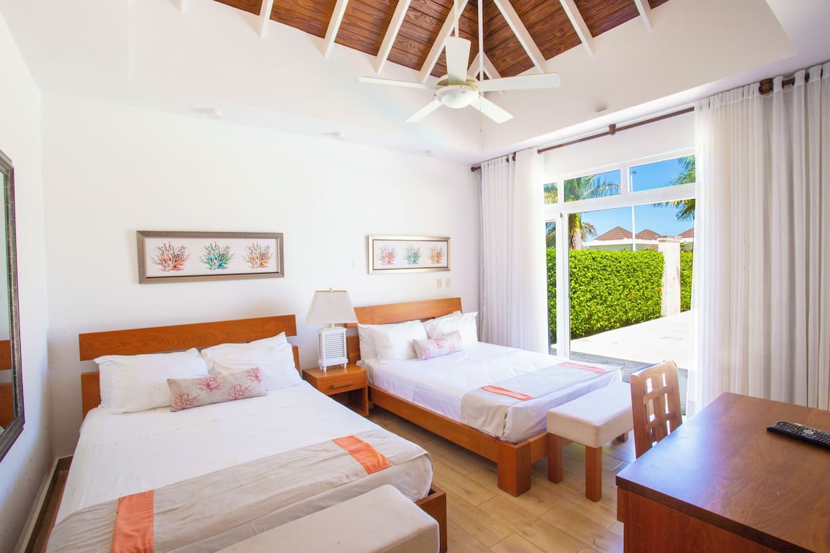A hotel room with two beds, a wooden ceiling, a ceiling fan, a window with curtains, and a table with a remote control.