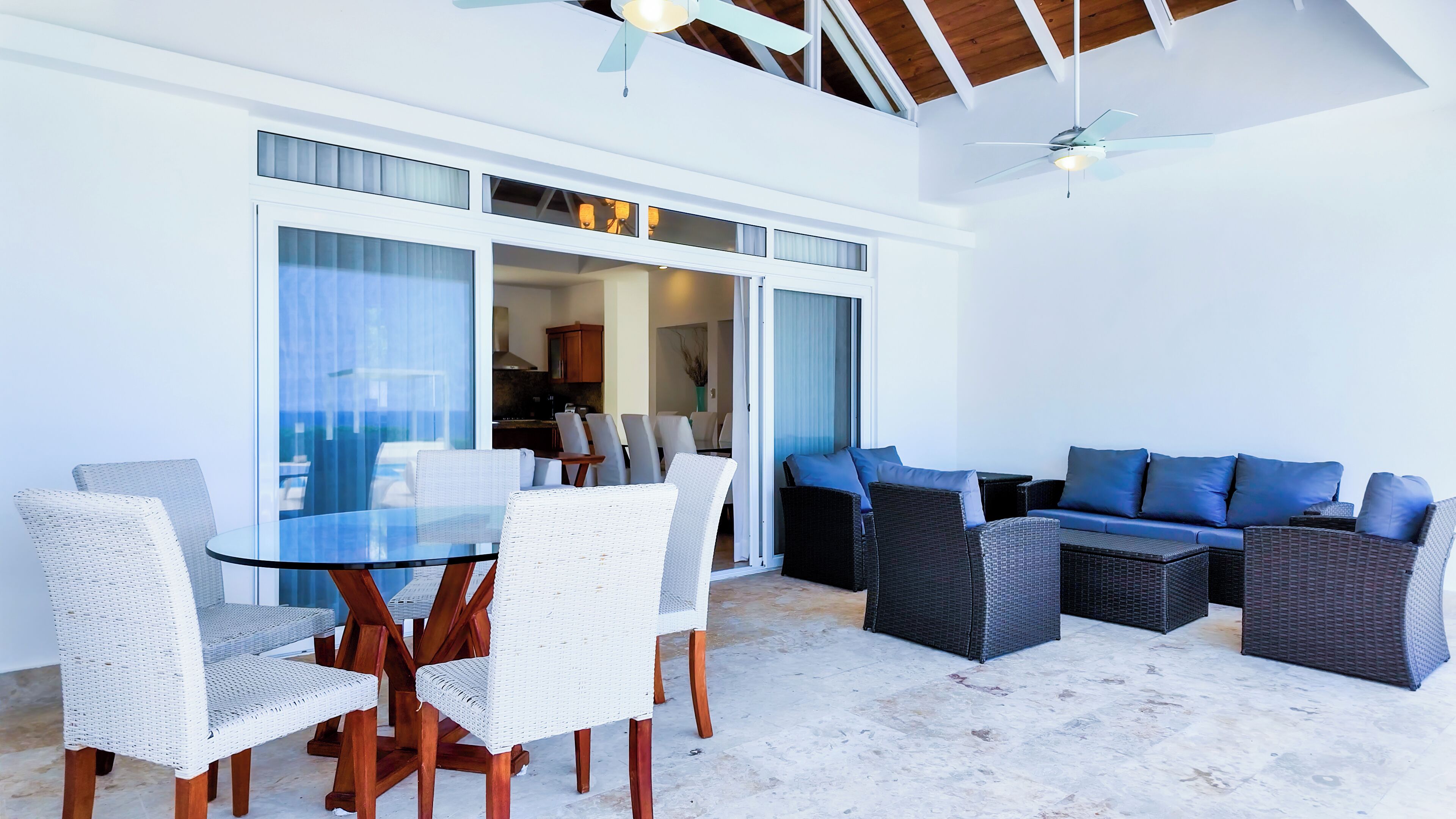 A modern outdoor dining area with a glass table and wicker chairs, adjacent to a blue sofa and wicker lounge chairs.