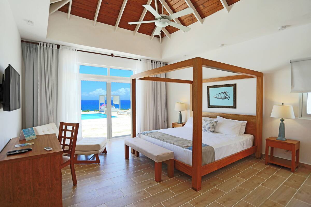 A four-poster bed with a view of the sea, a desk with a chair, and a small bedside table with a lamp.