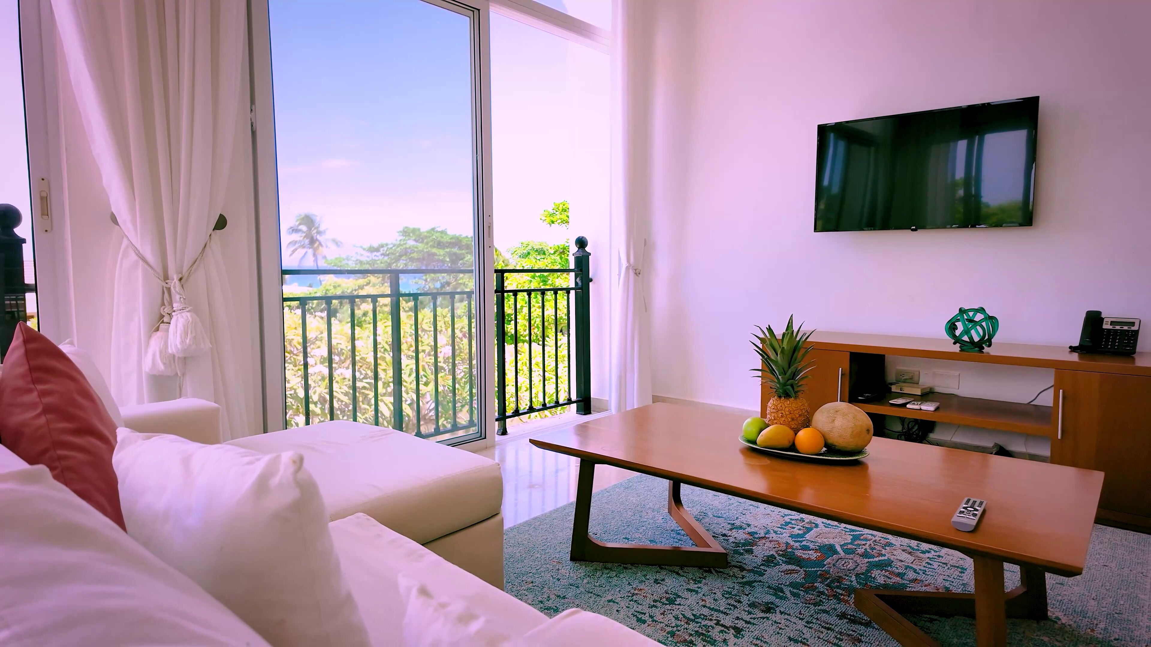 A hotel room with a balcony, a sofa, a coffee table with fruits, a TV, and a desk with a phone.