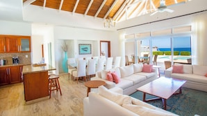 A spacious living area with a large sofa, a coffee table, and a dining area with chairs and a table. There is a kitchen area with wooden cabinets and a bar counter. The room has large windows with a view of the ocean. - Santa Fe Club (Sosúa)
