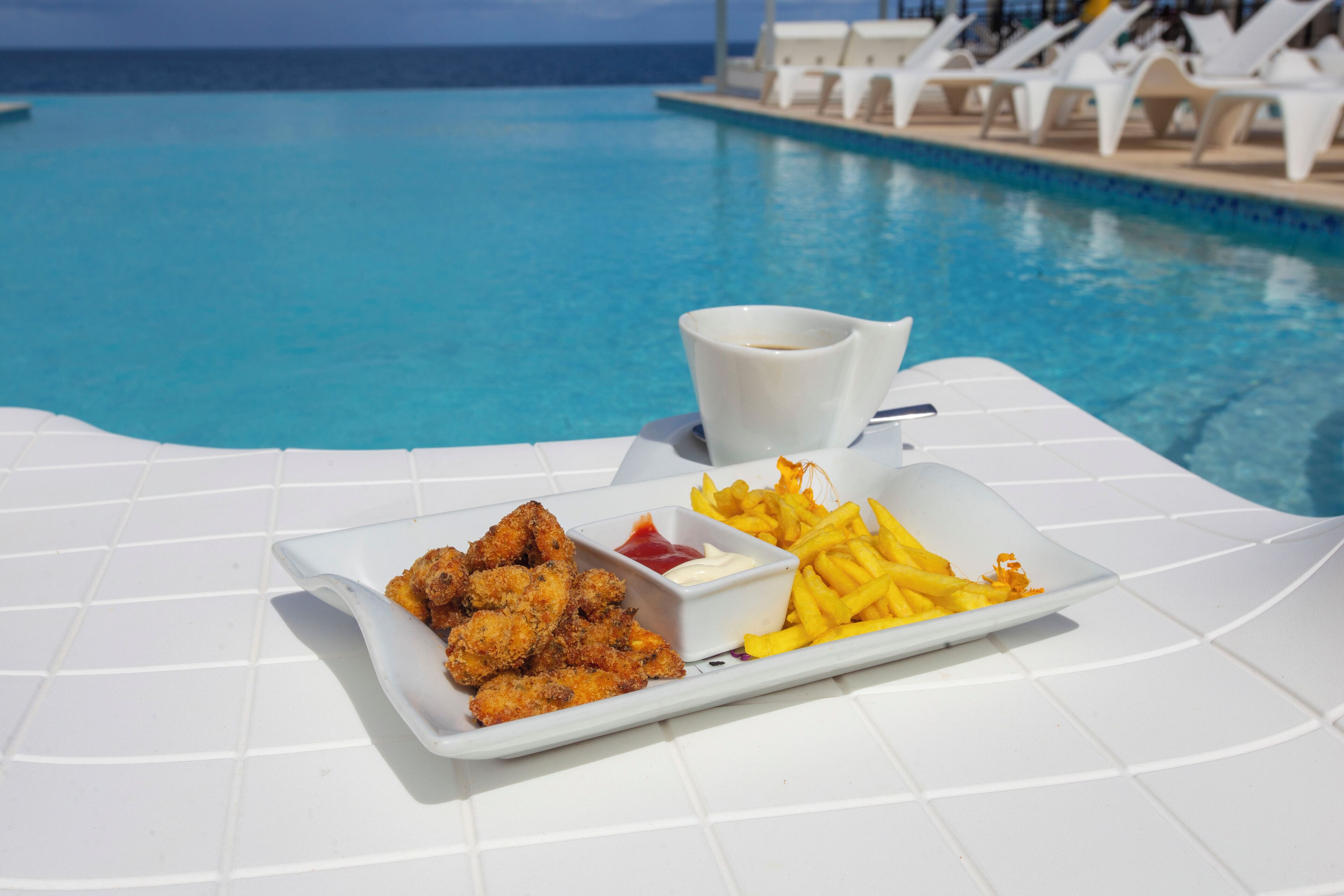 Ein Esstisch mit gebratenem Hähnchen, Pommes Frites, einer Kaffeetasse und Blick auf ein Schwimmbecken mit Liegestühlen.