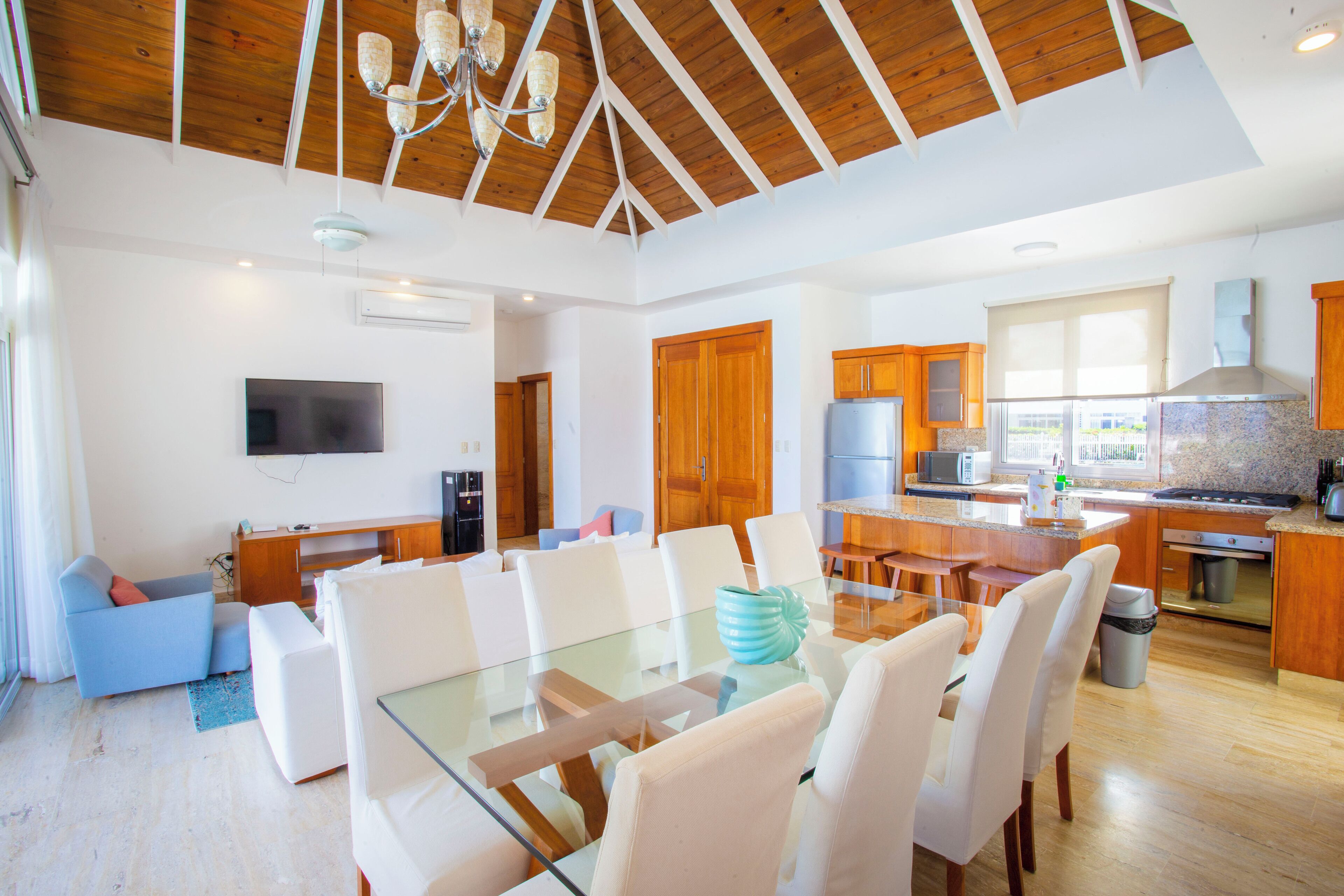 Un salón amplio con una mesa de vidrio rodeada de sillas blancas, techo de madera y zona de cocina con armarios de madera y un ventanal grande.