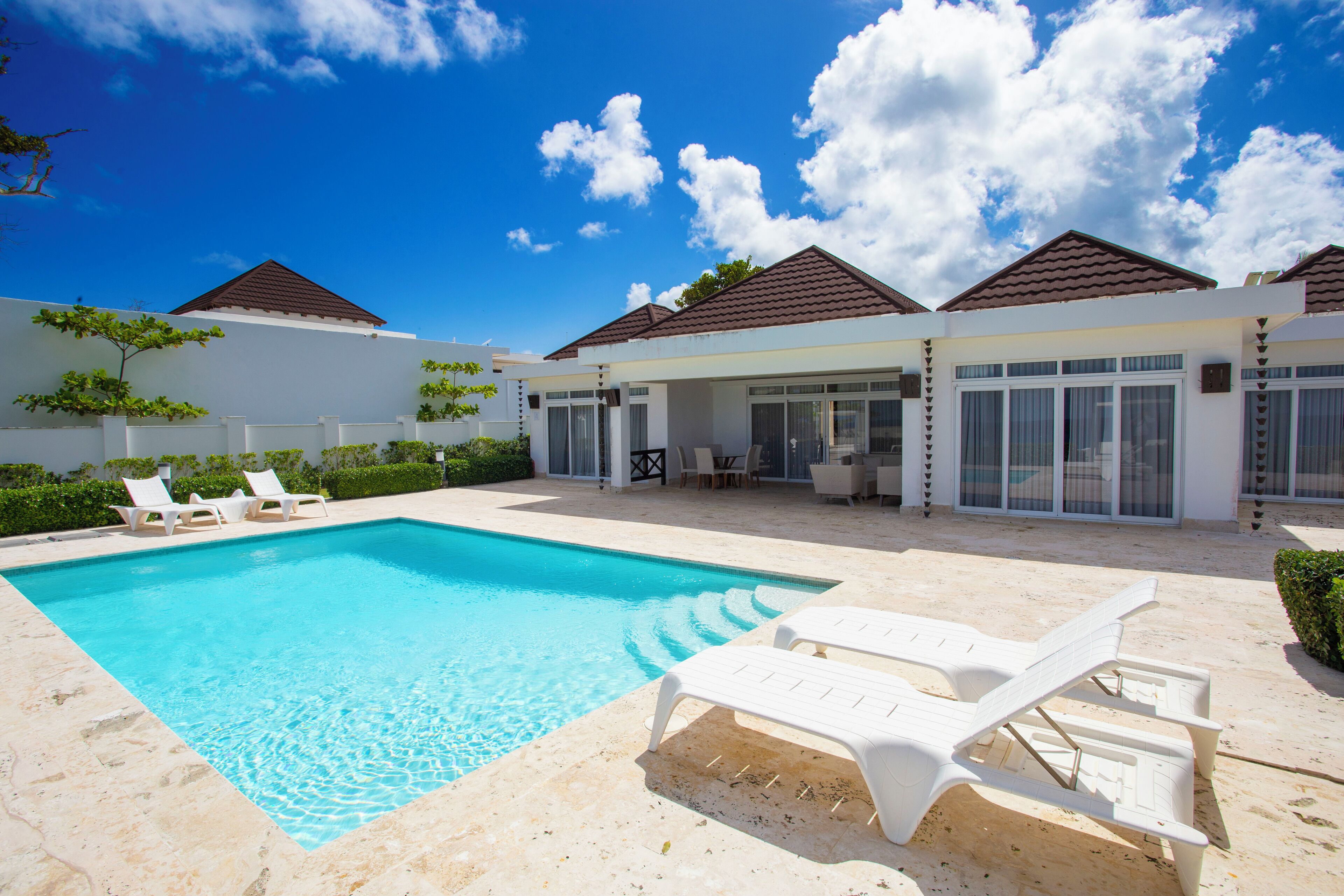A swimming pool with lounge chairs, a covered patio, and a white building with a tiled roof.
