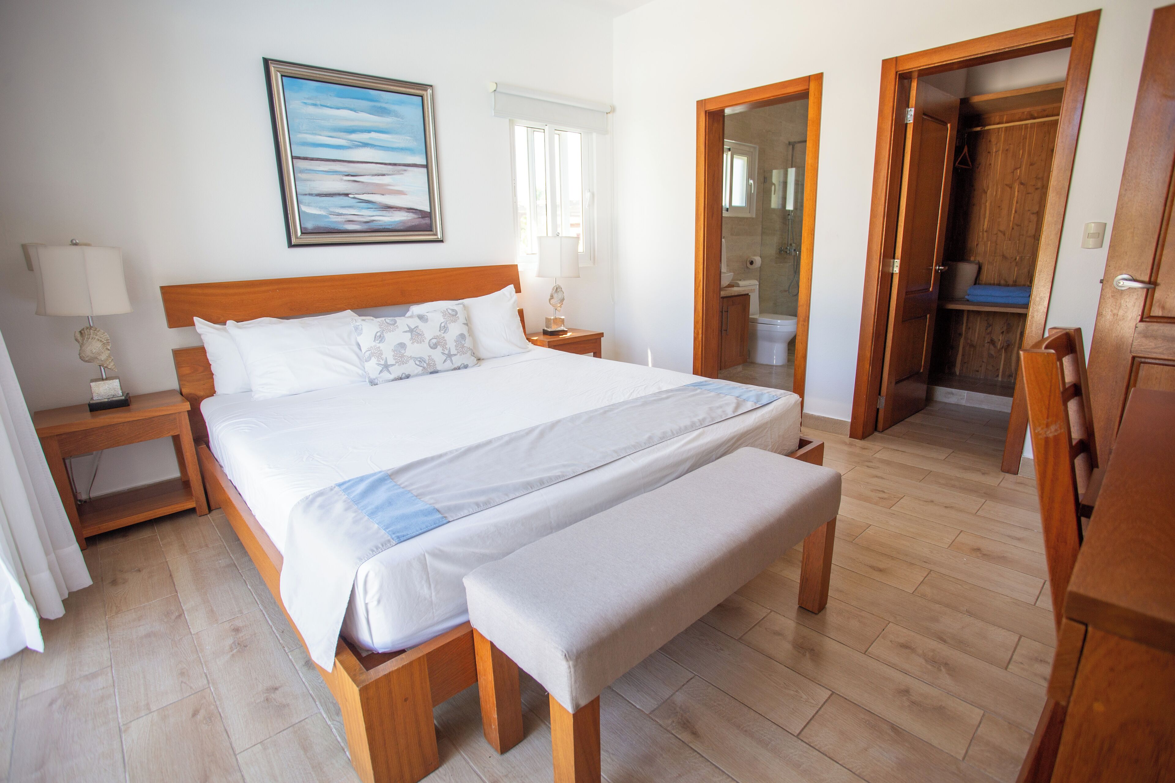 Une chambre à coucher avec un grand lit, des tables de chevet, un banc, une armoire en bois et une salle de bain visible à travers une porte ouverte.