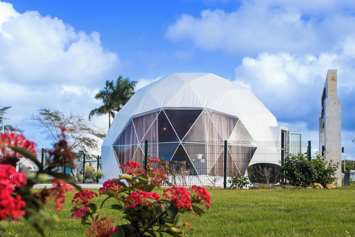 A geodesic dome structure with a transparent section, surrounded by a fence, and set in a grassy area with flowers.
