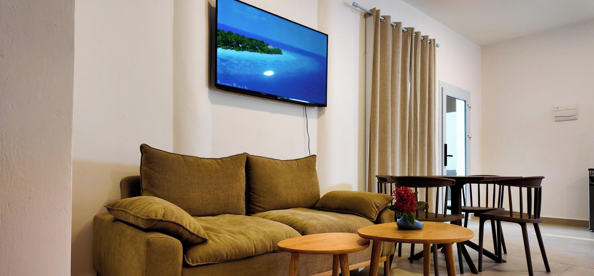 Un salon comprenant un canapé, un téléviseur fixé au mur, une table à manger avec des chaises et une fenêtre avec des rideaux.