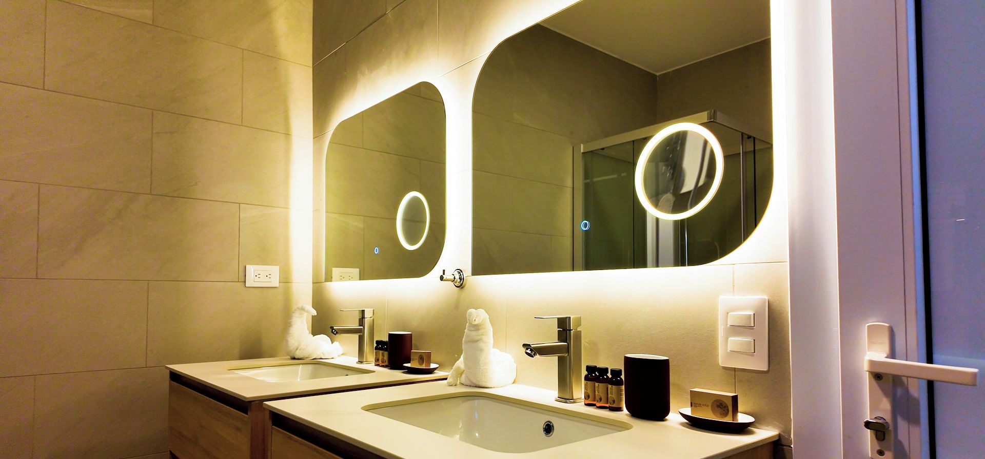 Une salle de bain moderne avec deux vasques, un miroir doté d’un éclairage circulaire et un porte-serviettes.