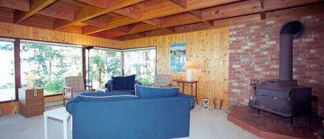 A cozy cabin interior with a blue sofa, wooden ceiling beams, a brick fireplace, and large windows offering a view of the outdoors.