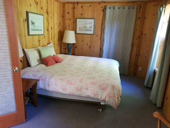 A bedroom with wooden panel walls, a bed with floral bedding, two pillows, a nightstand, a lamp, and a window with curtains. - Captain s Cottage (Coupeville)