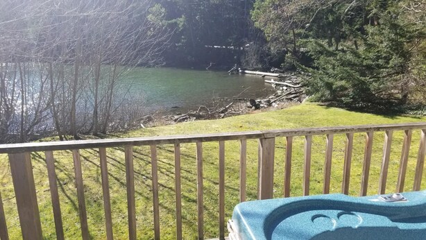 A wooden deck with a hot tub, overlooking a lake with trees and a grassy area.