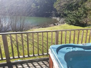 Un terrazzo in legno con una vasca idromassaggio, affacciato su un lago circondato da alberi e una zona verde.