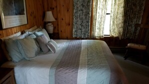 A neatly made bed with a quilted bedspread, a wooden nightstand with a lamp, and a chair in a room with wooden walls and floral curtains.