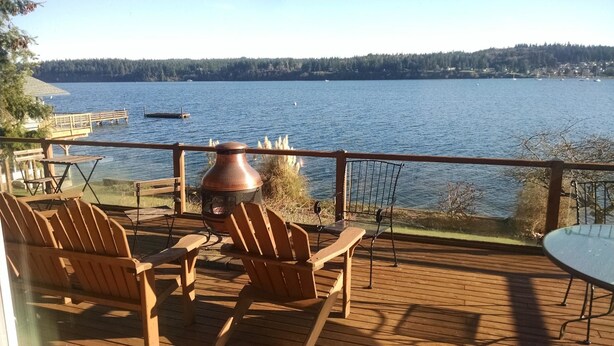 Terraza de madera con mobiliario exterior y un gran hogar de piedra, con vista a un lago con barcos y una orilla arbolada.