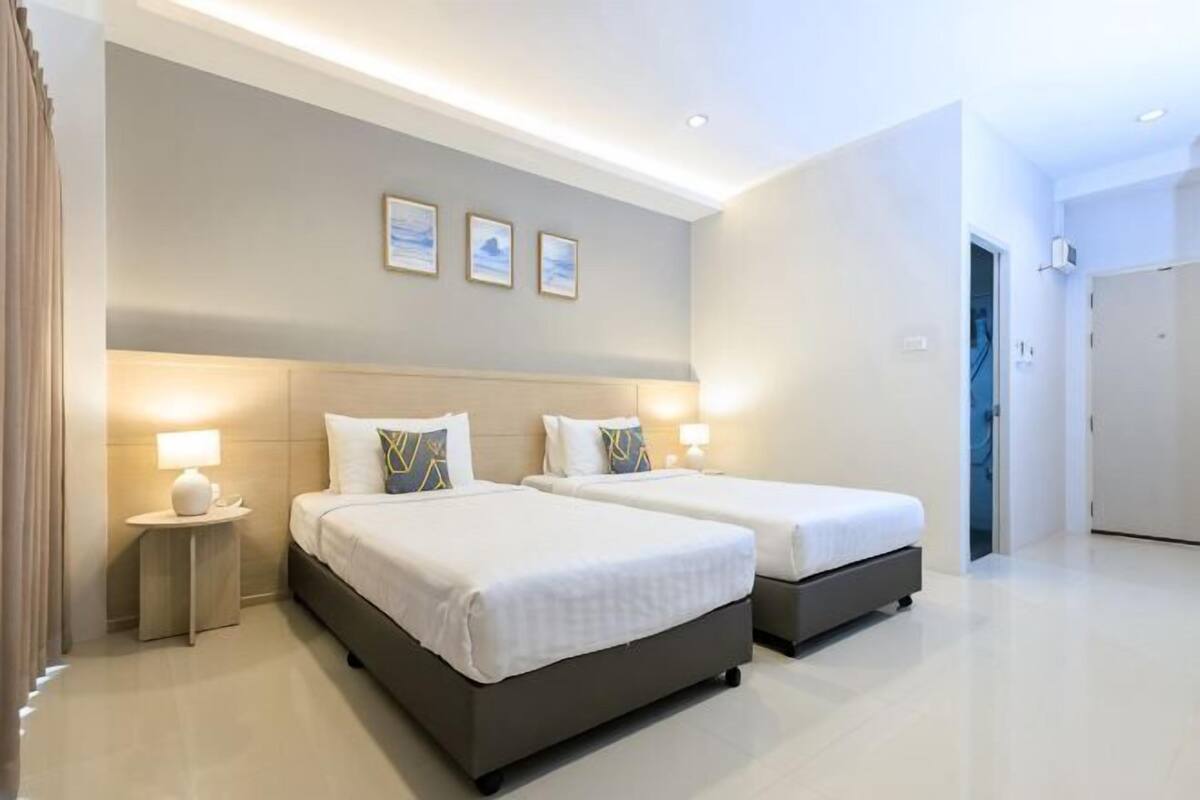 Habitación de hotel con dos camas, una mesita de noche, una lámpara de pared y una puerta que da a otra habitación.