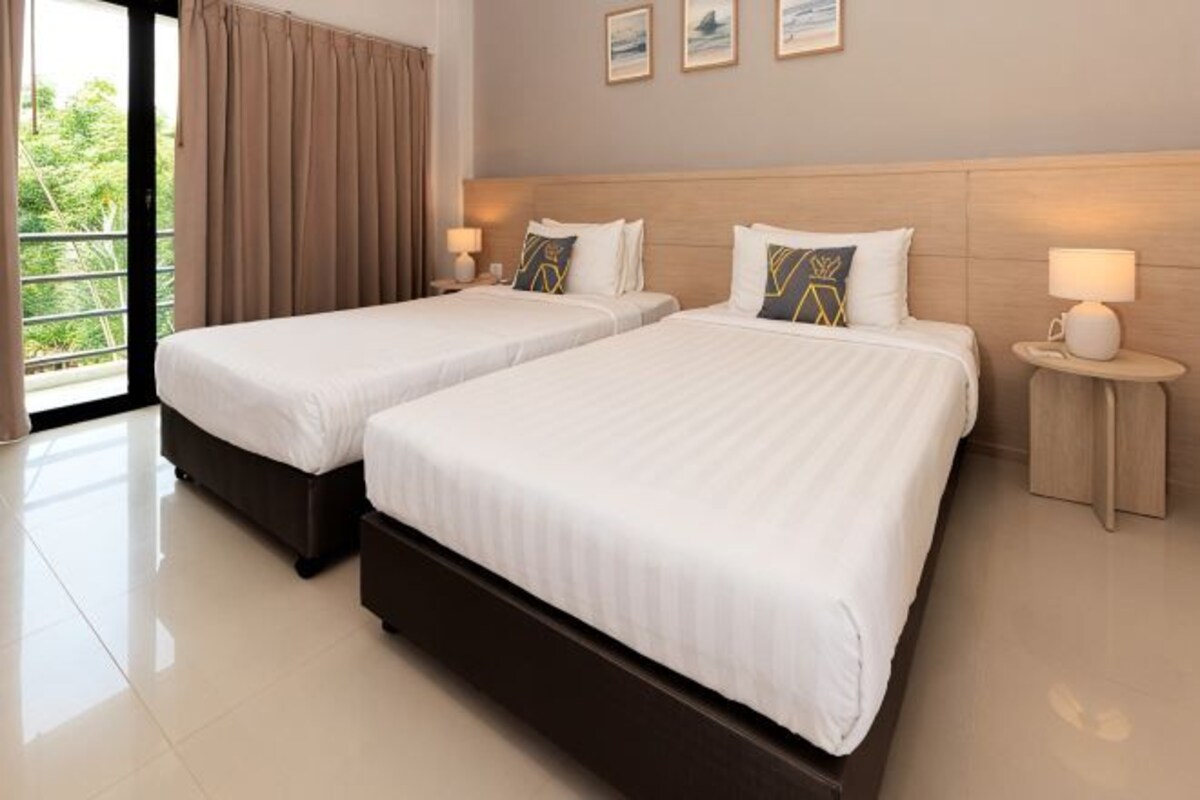 Habitación de hotel con dos camas, un ventanal grande con cortinas, un cabecero de madera y una mesita de noche con lámpara.