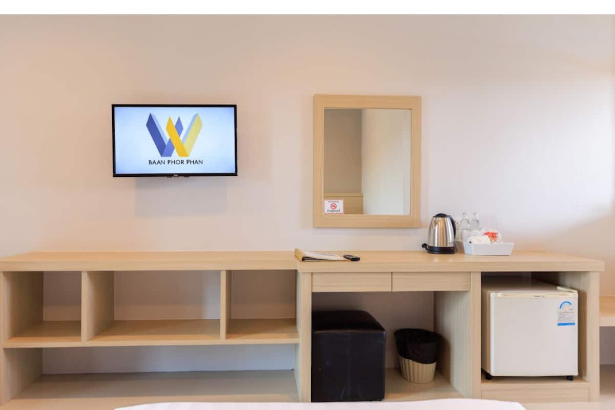 Una habitación de hotel con un escritorio de madera, un televisor mostrando 'BAAN PHOR PHAN', un espejo, un refrigerador y un cesto de basura.