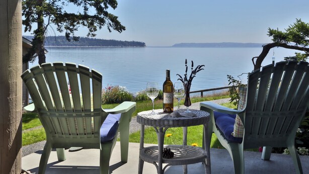 Two green Adirondack chairs facing a lake with a bottle of wine and glasses on a small table.