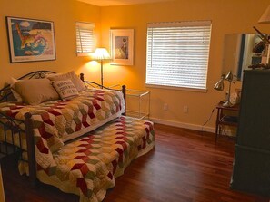A bedroom with a bed, a bedside table, a lamp, a window with blinds, and a painting on the wall.