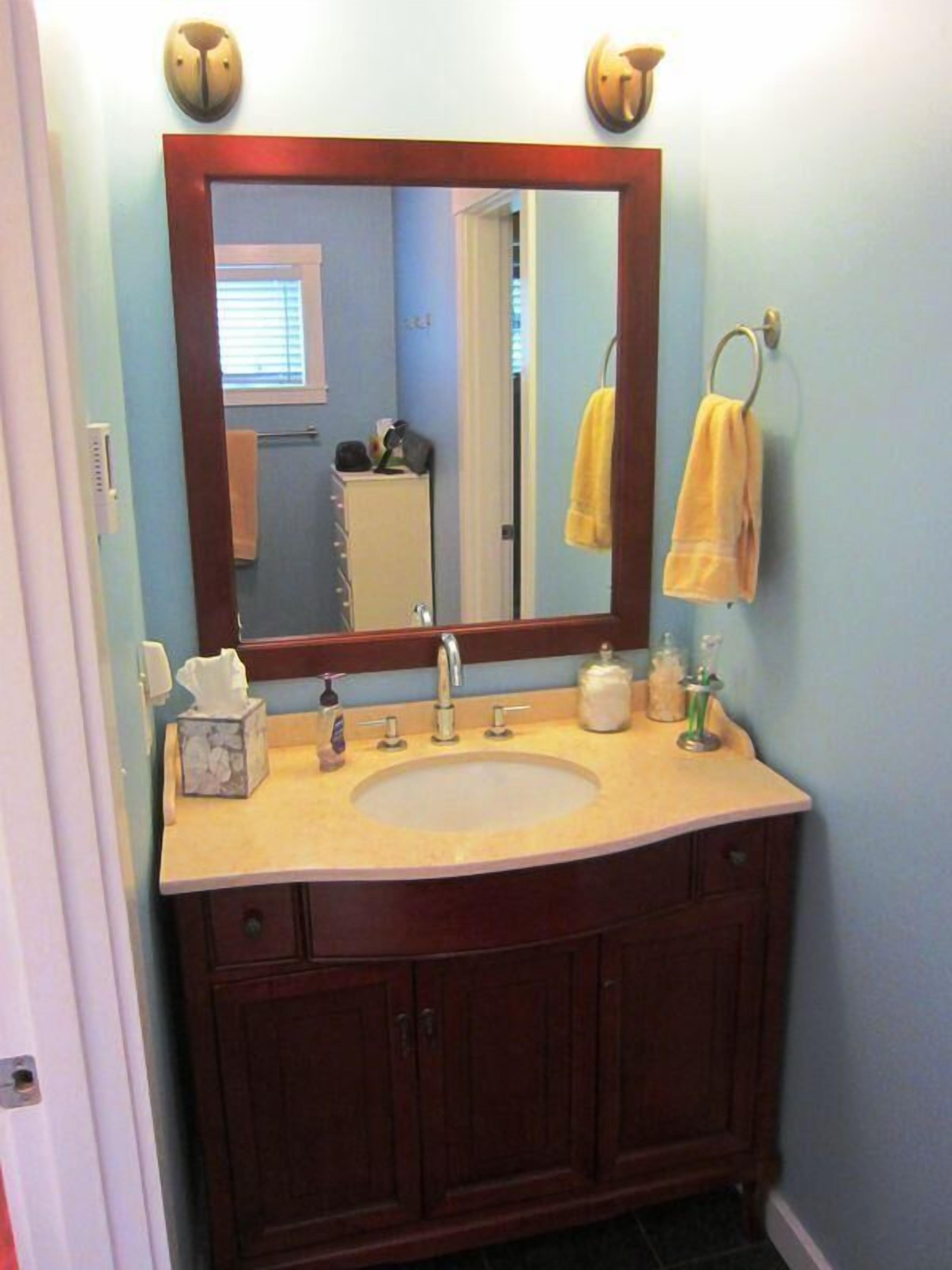 A bathroom with a wooden vanity, a sink, and a mirror. There are towels hanging on a rack, a soap dispenser, and a tissue box on the vanity.