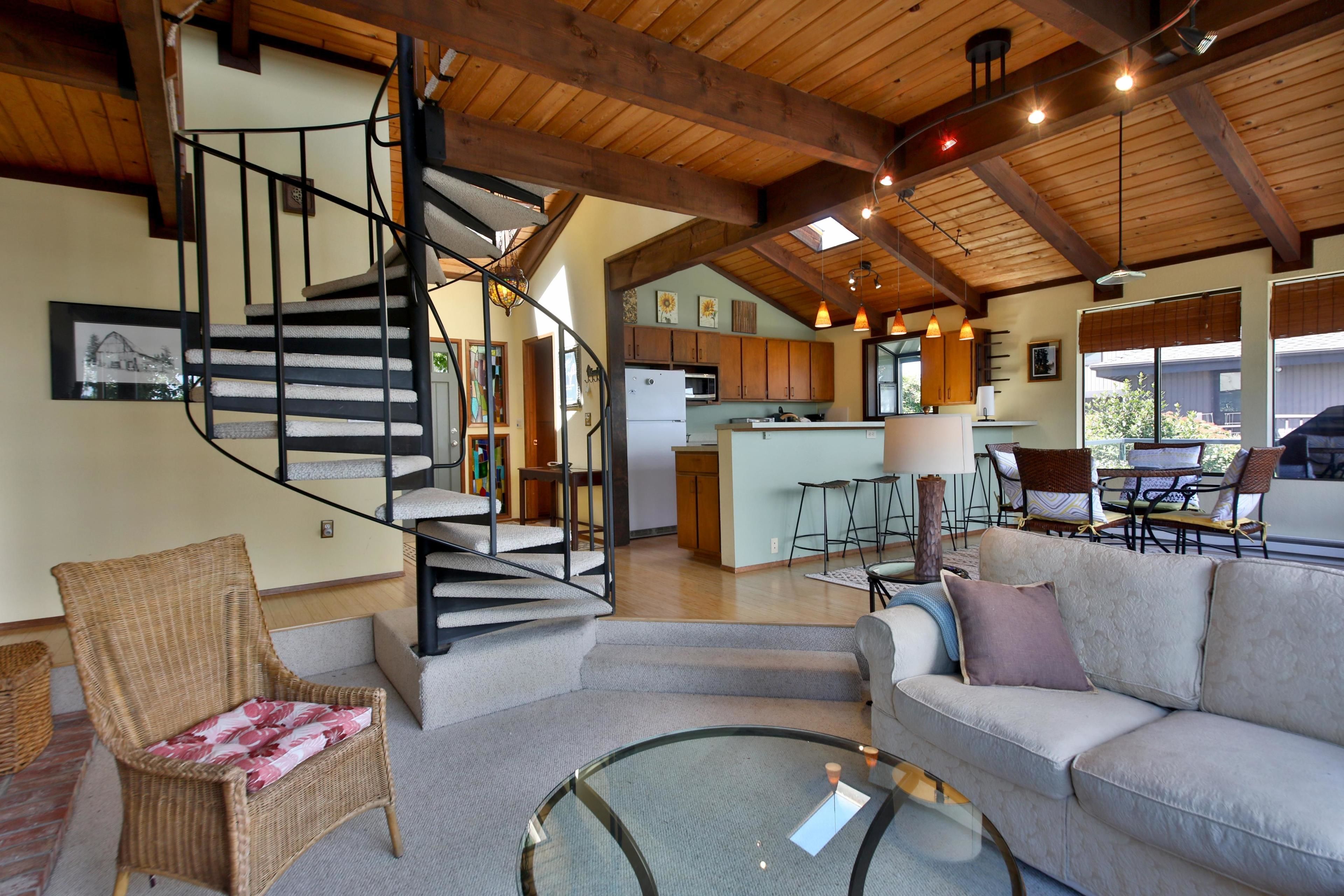 A spacious living area with a staircase, a sofa, a glass coffee table, and a kitchen in the background.
