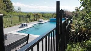 A swimming pool with a wooden deck and lounge chairs, surrounded by greenery and a view of the landscape. - Herb Cottage (Haumoana)