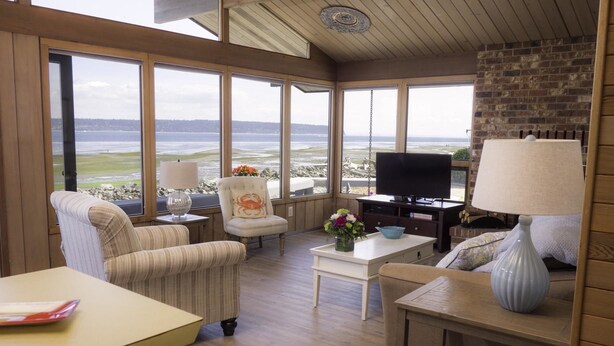 Una sala de estar acogedora con ventanas amplias que dan a una playa, un televisor y una chimenea.