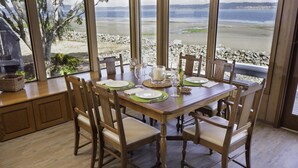 Mesa de comedor preparada para seis personas, con vista a una playa y un litoral rocoso.