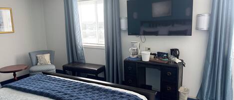 A hotel room with a bed, a desk with a coffee maker, a chair, a TV, and a window with curtains.