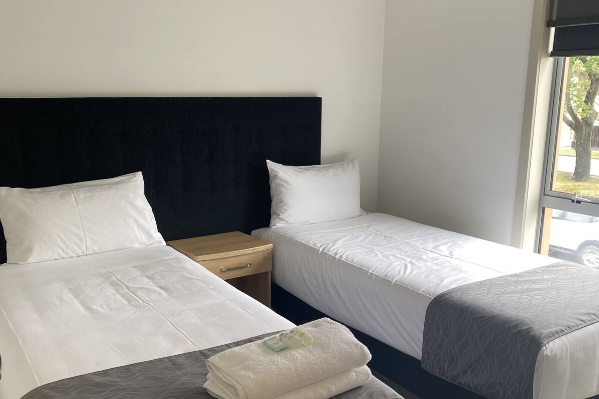 Ett hotellrum med två sängar, en mörk sänggavel, vita sängkläder och ett fönster med utsikt över träd.