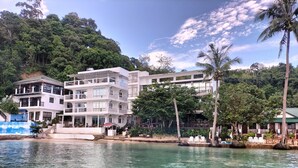 Un hotel de varios pisos con balcones, un área de piscina y exuberante vegetación al fondo.