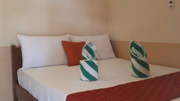 Quarto de hotel com cama, roupa de cama branca, manta vermelha e duas toalhas listradas em verde e branco.