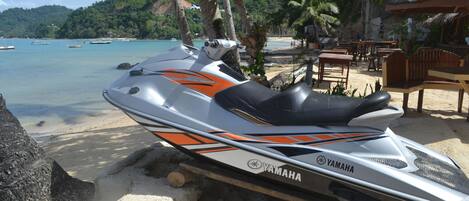 Een Yamaha jetski geparkeerd op een zandstrand, met uitzicht op de oceaan en een rieten hutje op de achtergrond.