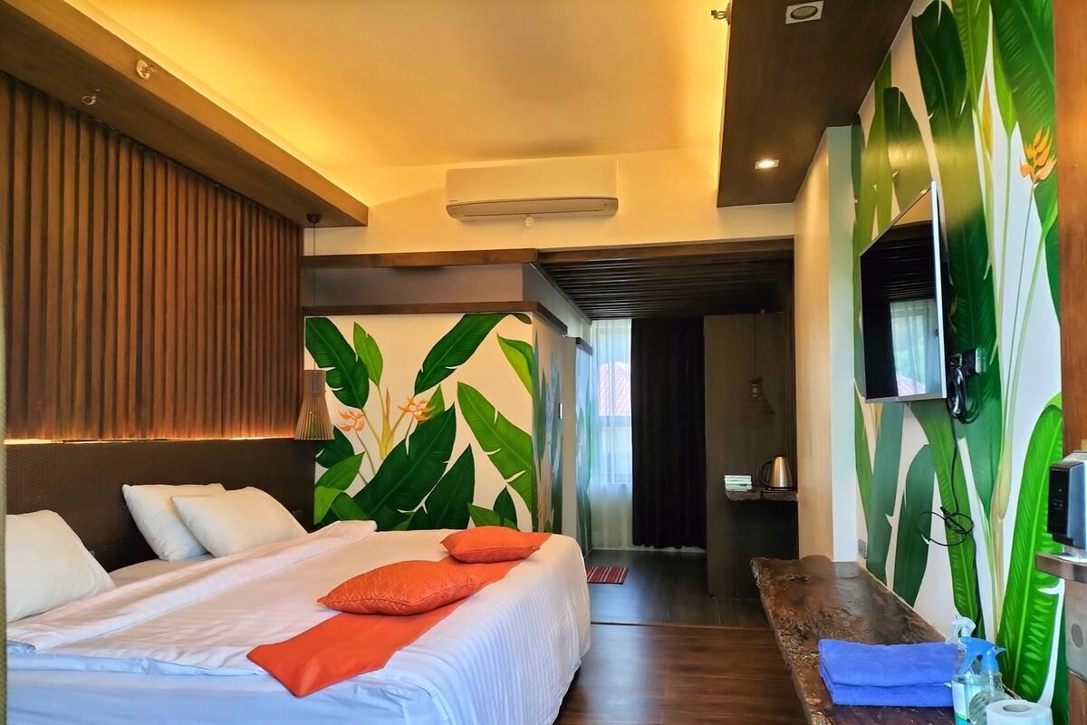 Ein Hotelzimmer mit Bett, Holz-Kopfteil, Wandmalerei mit tropischen Blättern, einem an der Wand montierten Fernseher und einer Holzbank mit Pflegeprodukten.