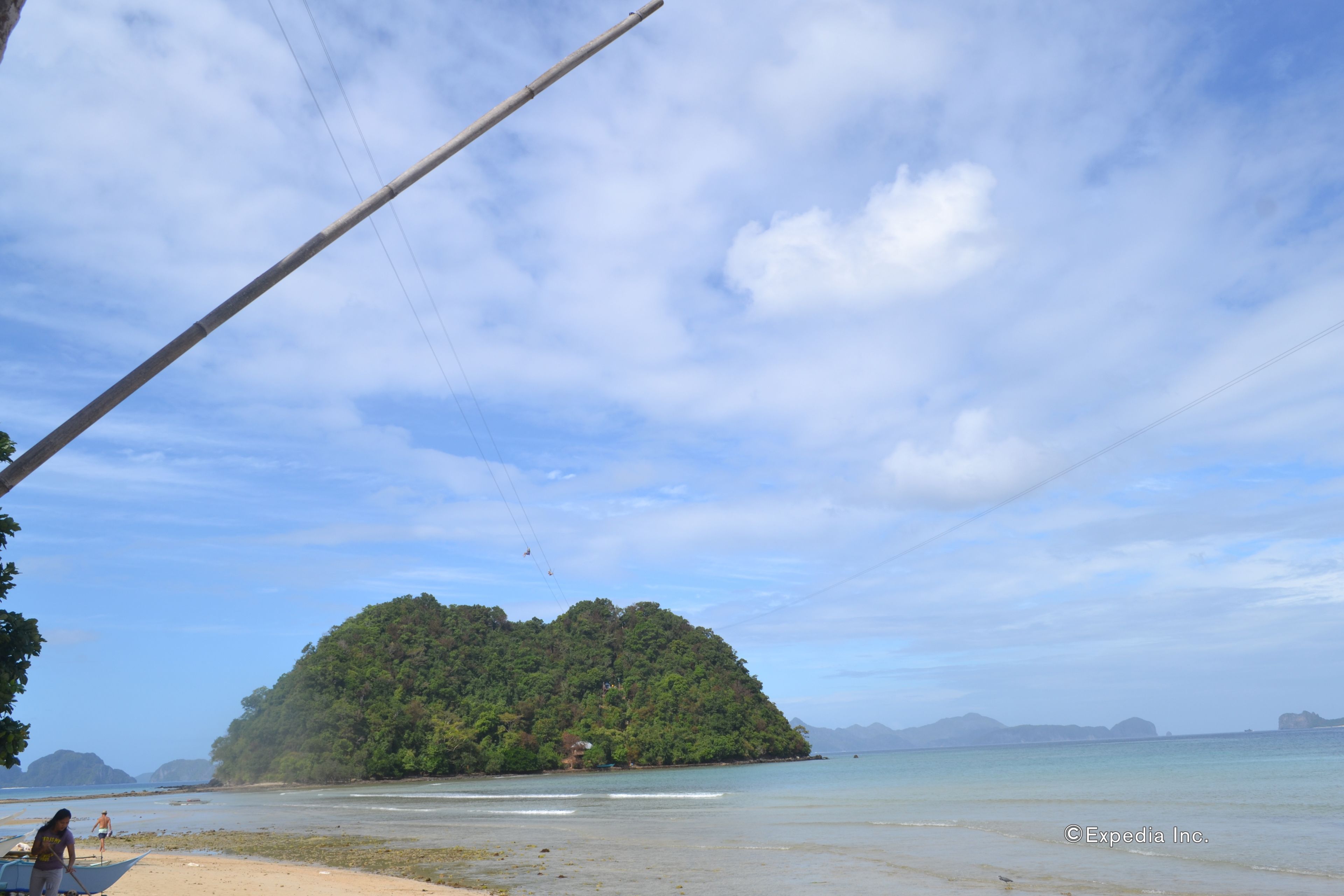Una playa con una pequeña isla en la distancia, cielos despejados y algunas personas caminando.