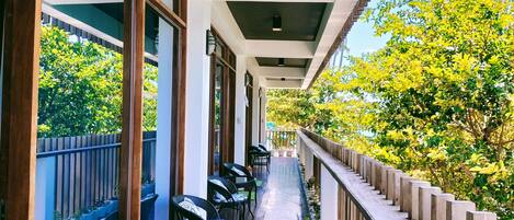 A covered outdoor corridor with wicker chairs and potted plants, leading to a balcony with a view of trees.