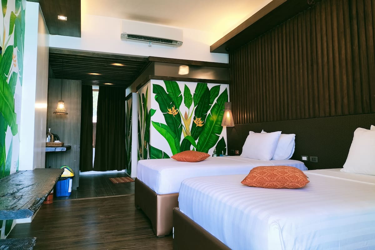 Ein Hotelzimmer mit zwei Betten, einer Holzbank, einer Wand mit einem tropischen Blatt-Wandbild und einem Deckenventilator.