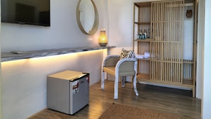 Una habitación moderna con televisión de pantalla plana, un espejo redondo, un refrigerador pequeño, sillas de mimbre, un mueble de madera con estantes y una lámpara de pie encendida.