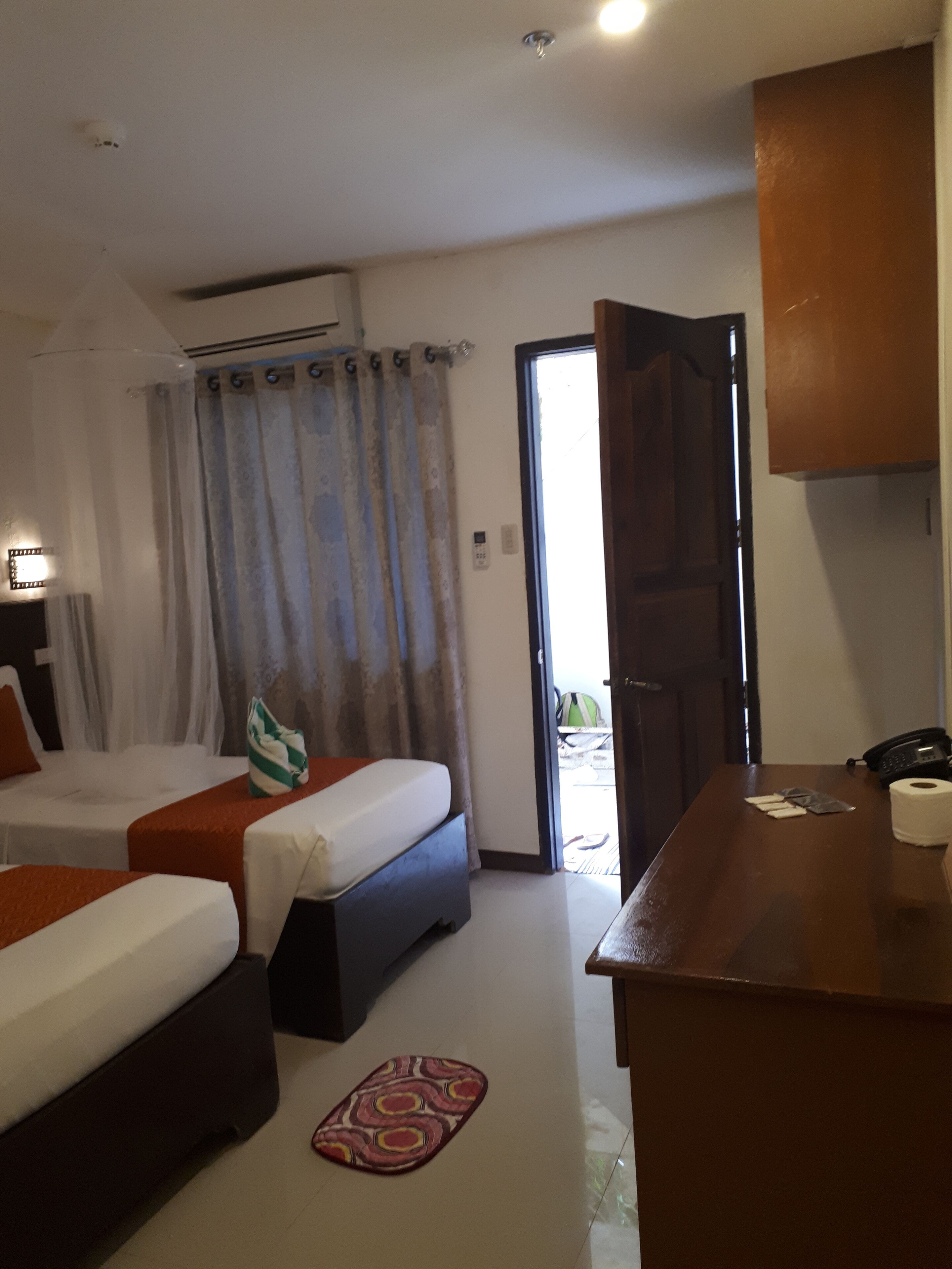 A hotel room with two beds, a small kitchenette, a bathroom, and a view of the outside.