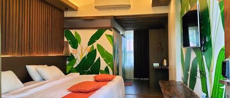 A hotel room with a bed, a wooden bench, a TV, and a wall with a tropical leaf mural.