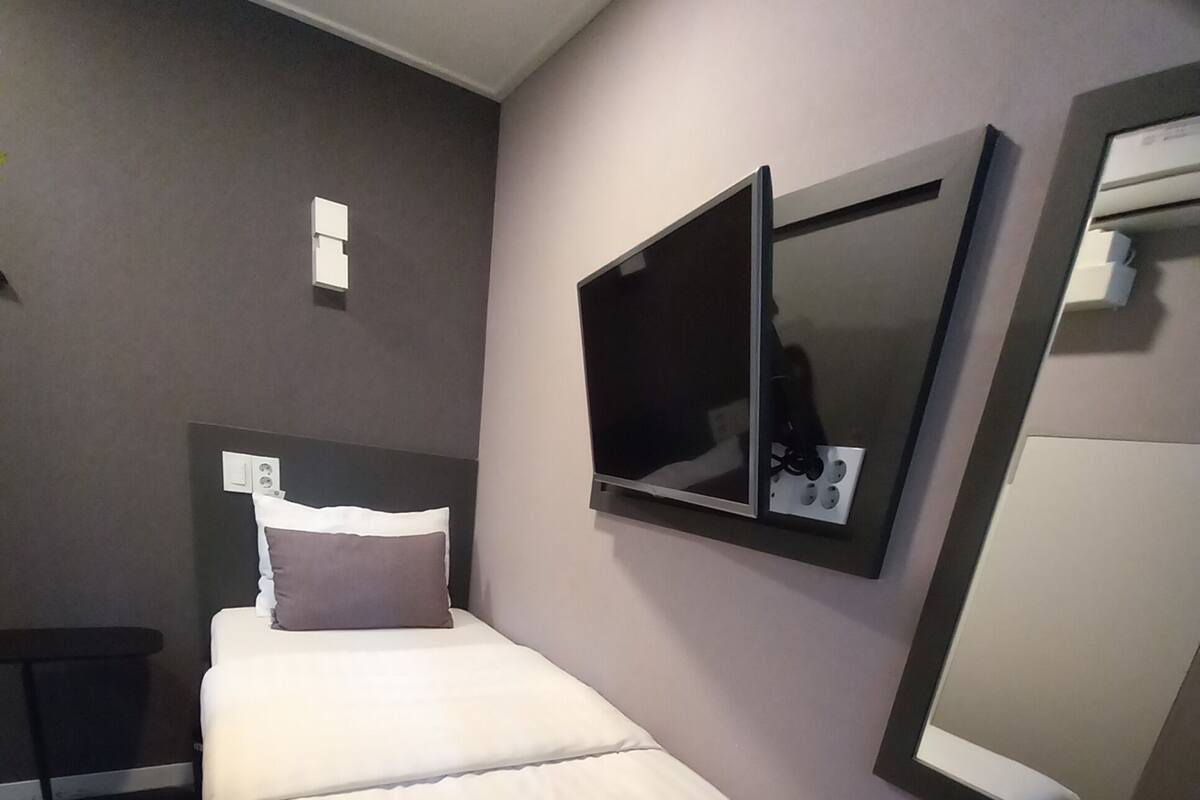 A hotel room with a bed, a TV mounted on the wall, a mirror, and a small table with a trash bin.