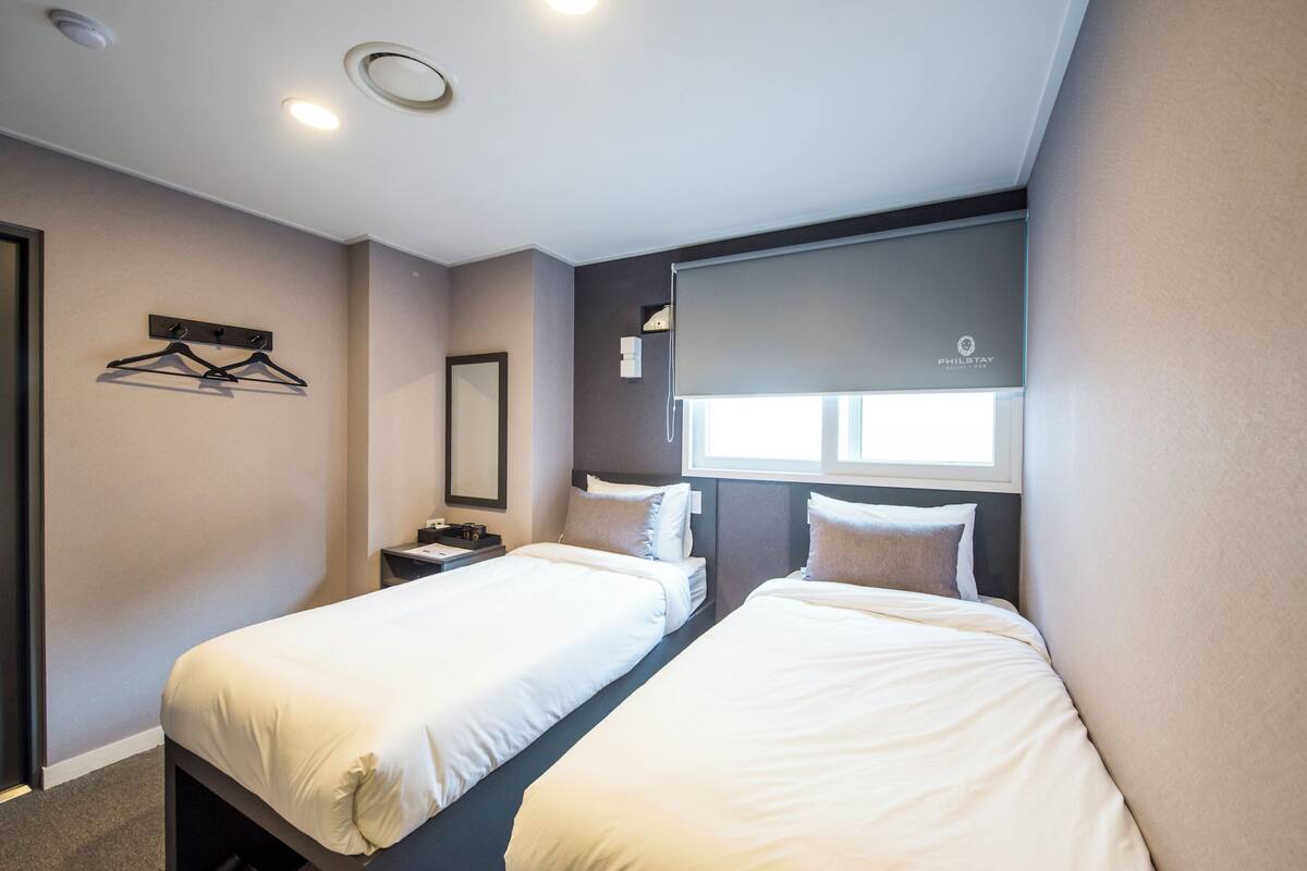 A hotel room with two single beds, a bedside table with a lamp, a wall-mounted shelf with hangers, a window with a blind, and a ceiling light fixture.