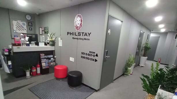 A hotel reception area with a reception desk, a clock, and a wall with room numbers and a logo. - Philstay Myeongdong Metro (Seoul)