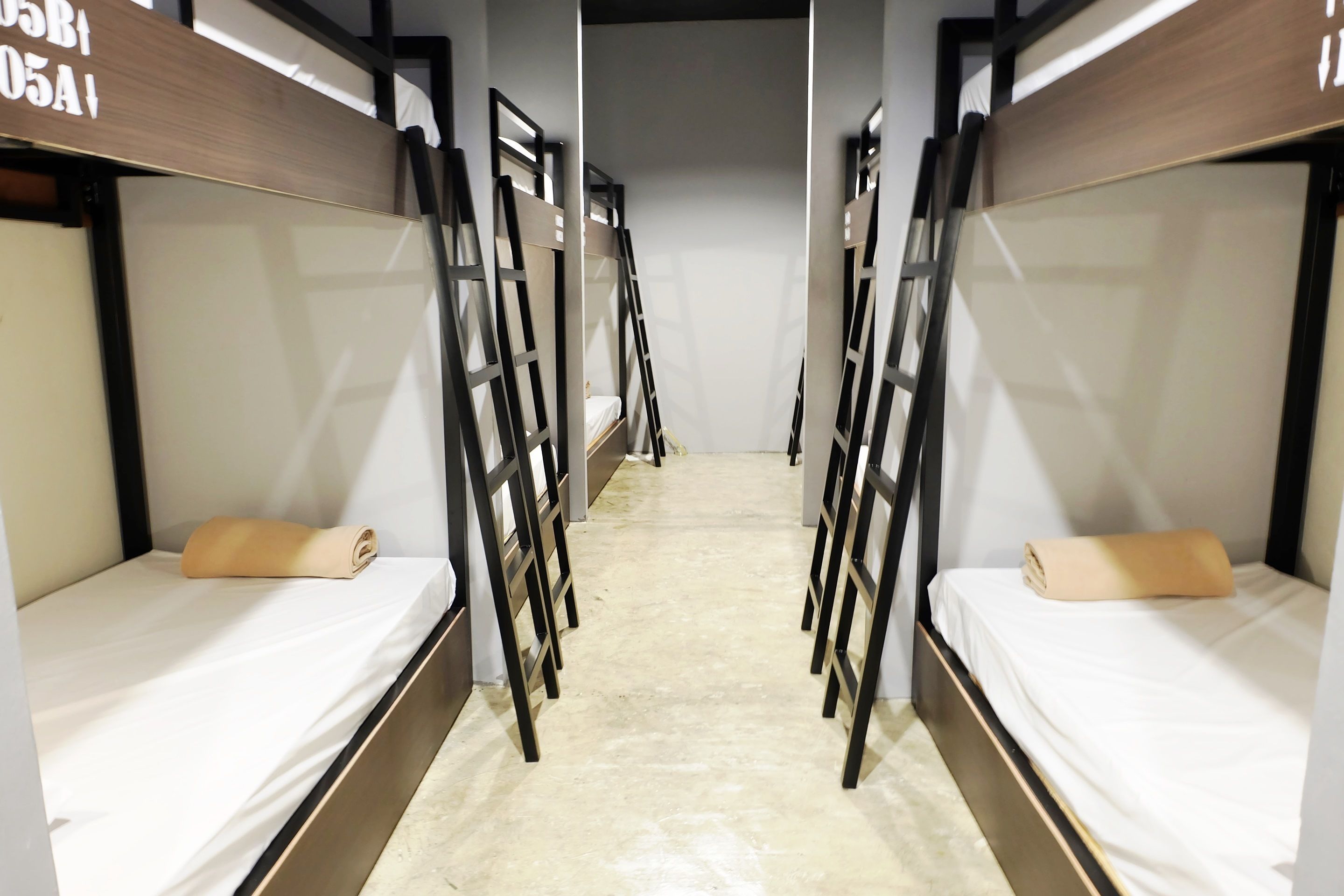 A corridor with bunk beds on either side, each with a mattress and a rolled-up blanket.
