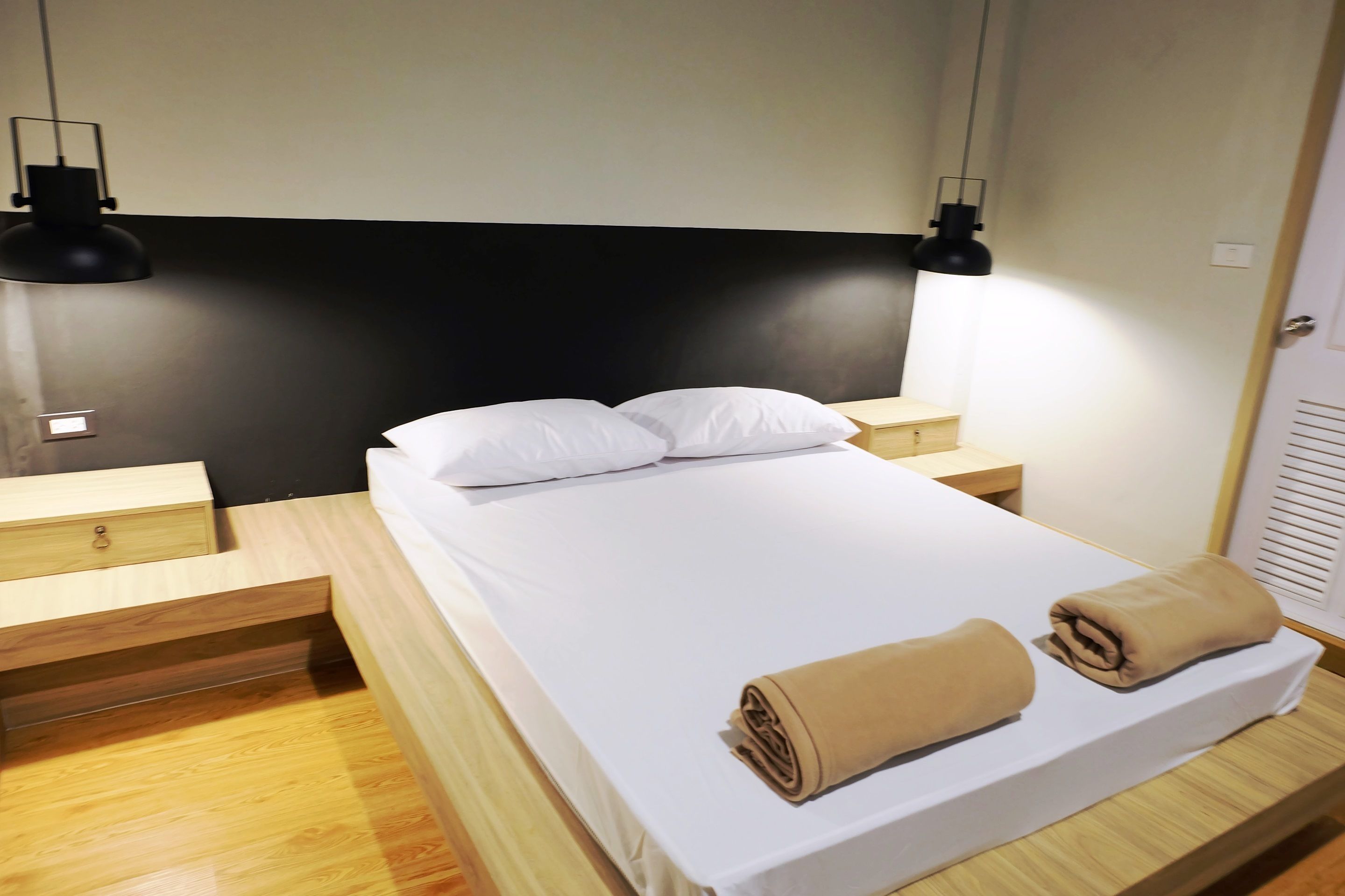 Een netjes opgemaakt bed met witte lakens en twee opgerolde handdoeken, houten nachtkastjes met lades, en zwarte hanglampen aan beide zijden van het bed.
