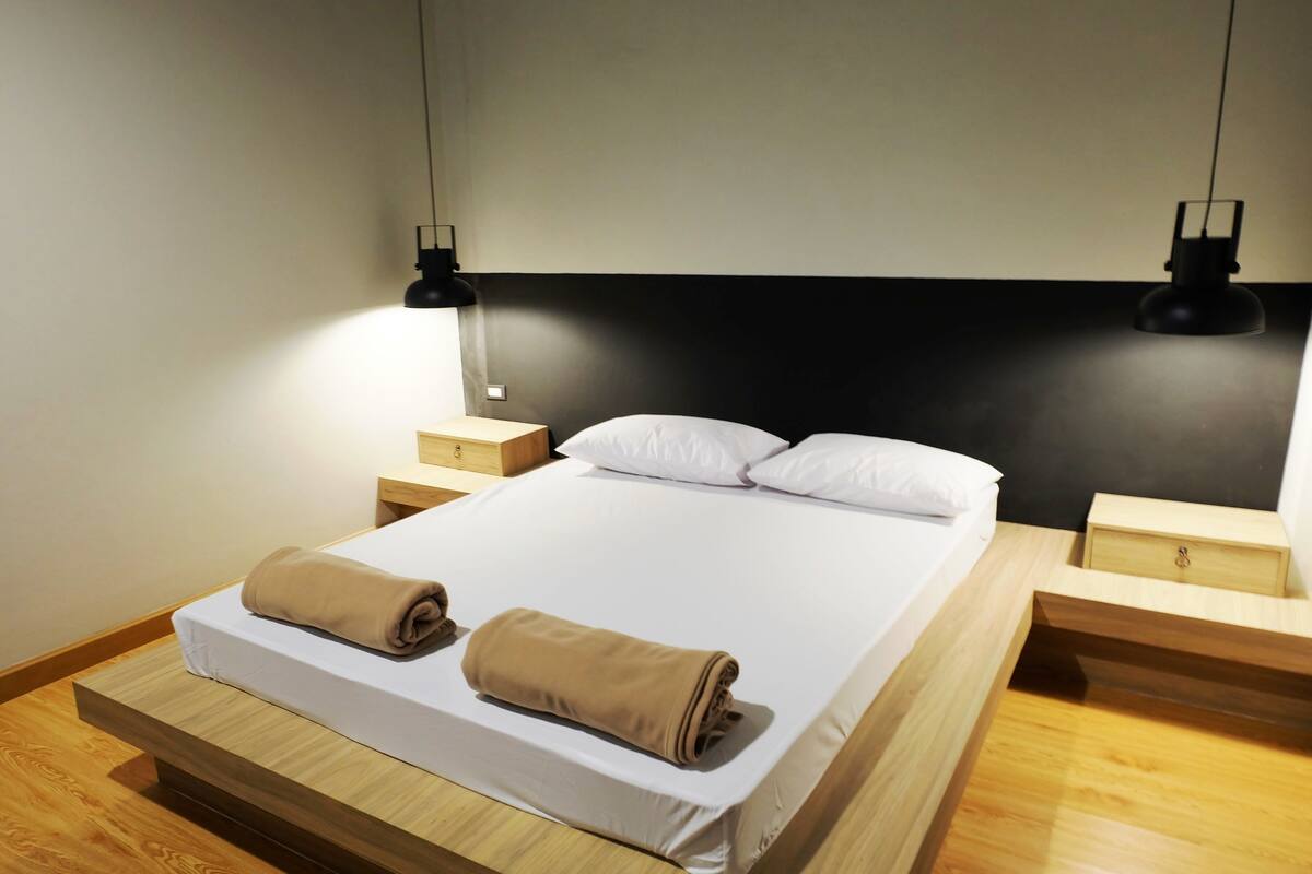 A neatly made bed with two rolled towels, wooden nightstands, and two pendant lights.