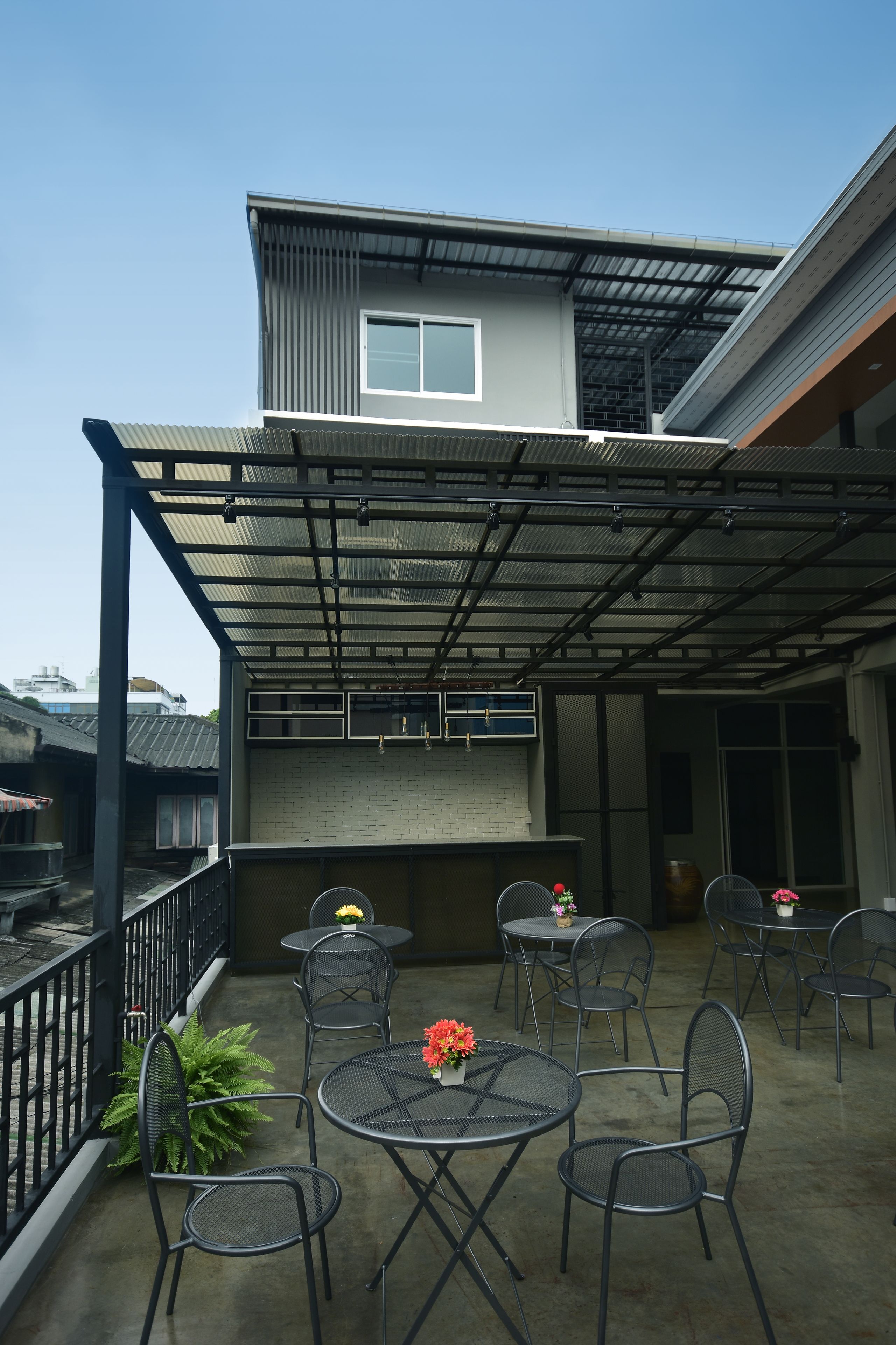 Un patio cubierto al aire libre con mesas y sillas de metal, un pequeño jardín y un edificio con techo y ventanas de metal.