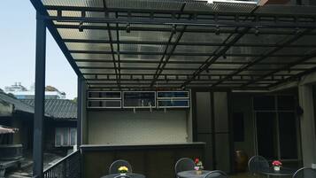 Un patio cubierto al aire libre con mesas y sillas de metal, un pequeño jardín y un edificio con techo y ventanas de metal.