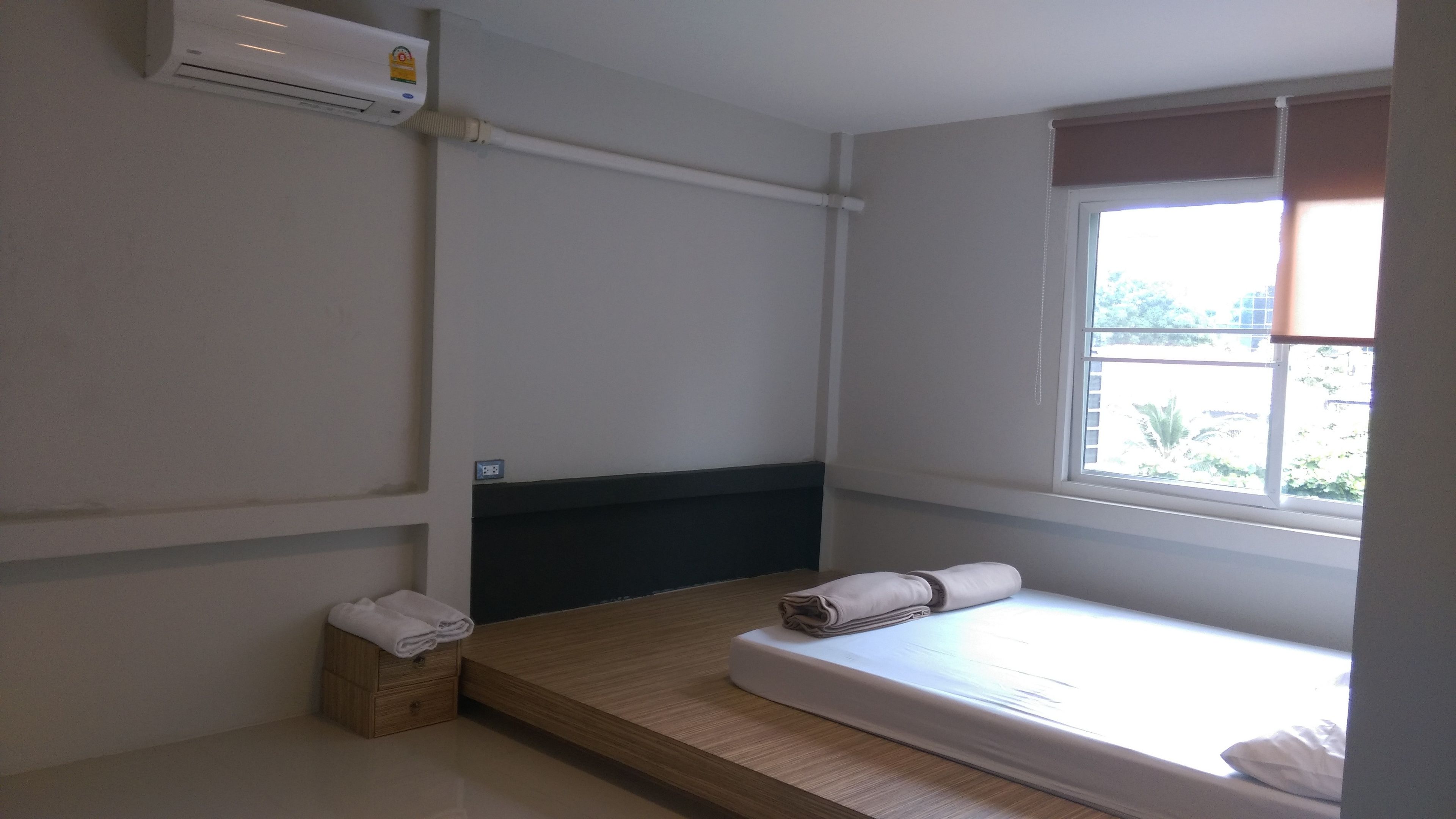 Ein kleines, sauberes Zimmer mit einem Einzelbett, einem Holz-Nachttisch, einer Wand-Klimaanlage und einem Fenster mit Blick nach draußen.
